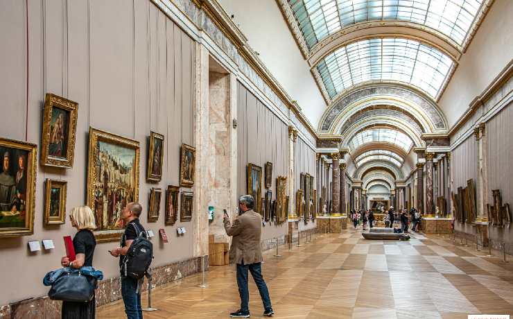 Vista di una galleria interna del museo Louvre con colori chiari e terrosi e tanti turisti che guardano e fotografano i quadri