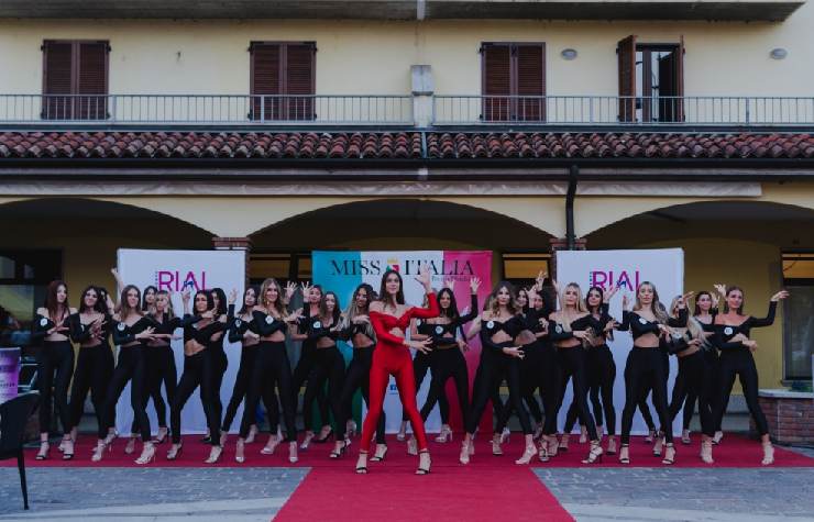 Tante ragazze partecipanti a Miss Italia in posa con al centro una ragazza vestita di rosso in un cortile